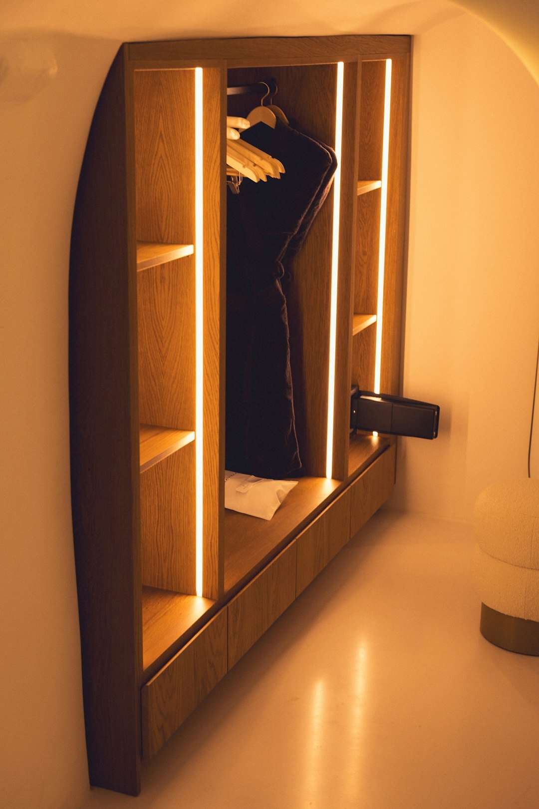 A cozy and modern wooden wardrobe illuminated by soft, warm lighting. The wardrobe features neatly arranged shelves and hangers, with a luxurious robe and a few neatly folded towels. The minimalist design and ambient lighting create a welcoming and comfortable atmosphere, ideal for a stylish bedroom or hotel room.