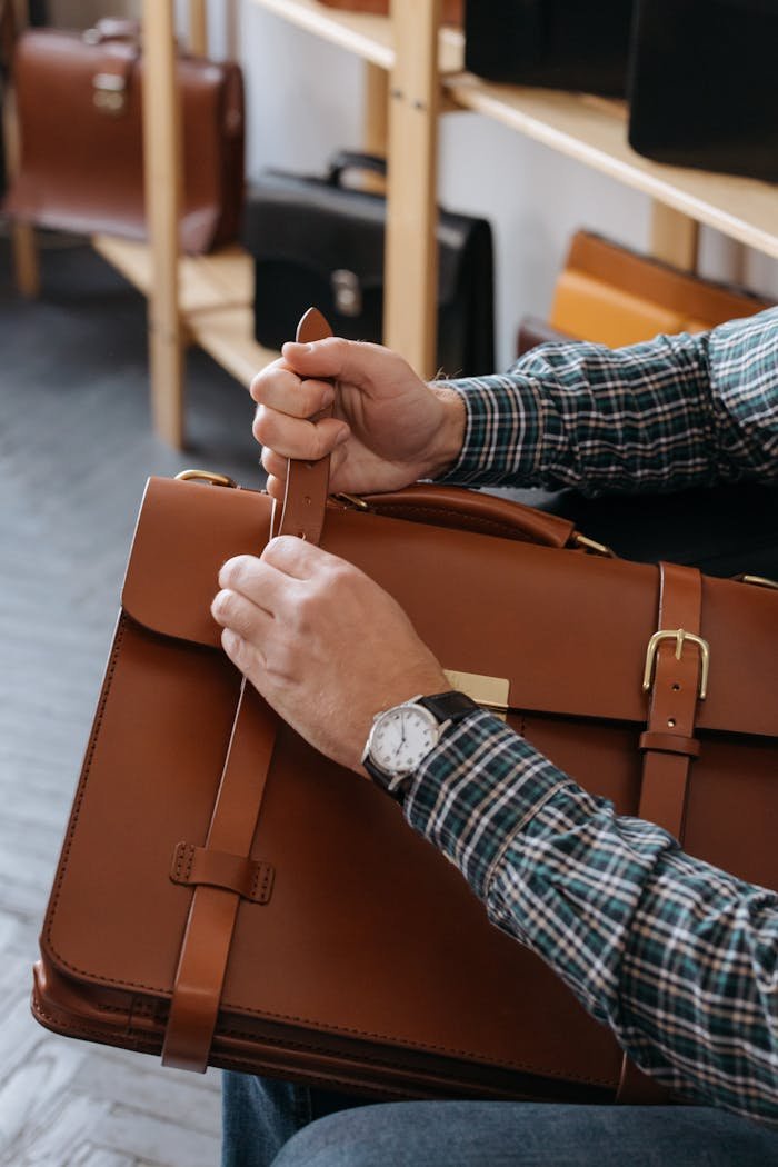A close-up shot of a mans hands adjusting a brown leather briefcase indoors.